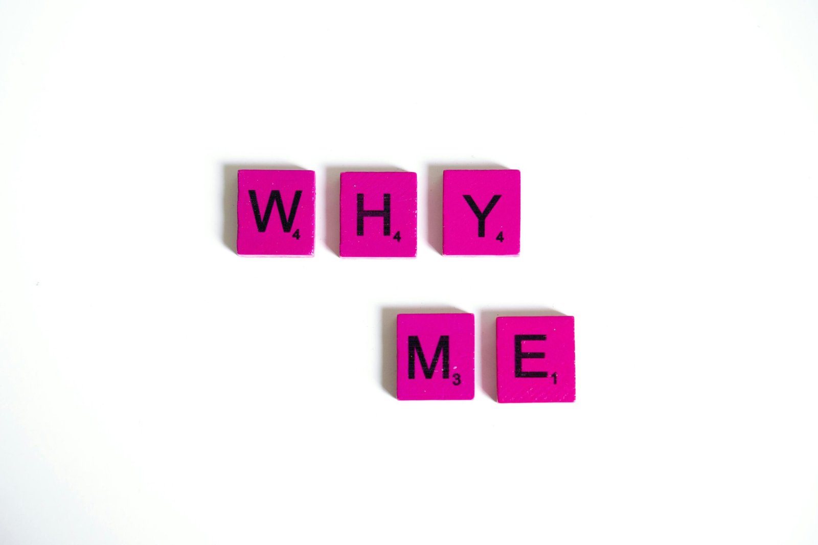 Pink Scrabble tiles form the words 'Why Me' on a white background, emphasizing questioning and introspection.
